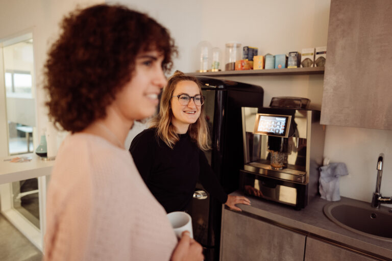 Bild von zwei Frauen in einer Küche an einem Kaffeeautomaten. Beide schauen in die selbe Richtung und lächeln.