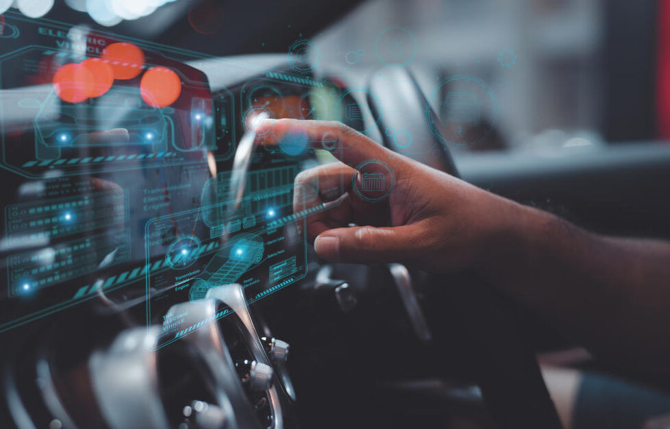 Ein Finger berührt eine virtuelle Anzeige am Armaturenbrett eines Autos, im Hintergrund ein Lenkrad