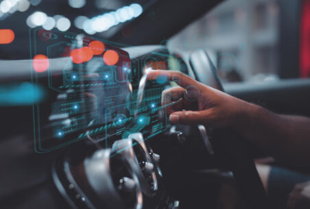 Ein Finger berührt eine virtuelle Anzeige am Armaturenbrett eines Autos, im Hintergrund ein Lenkrad
