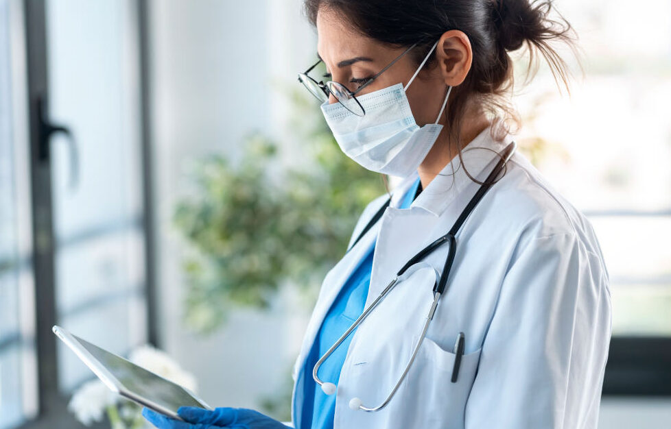 Person trägt Brille, Mundschutz, weiße Jacke, blaue Handschuhe, ein Stethoskop und in der Hand ein Tablet
