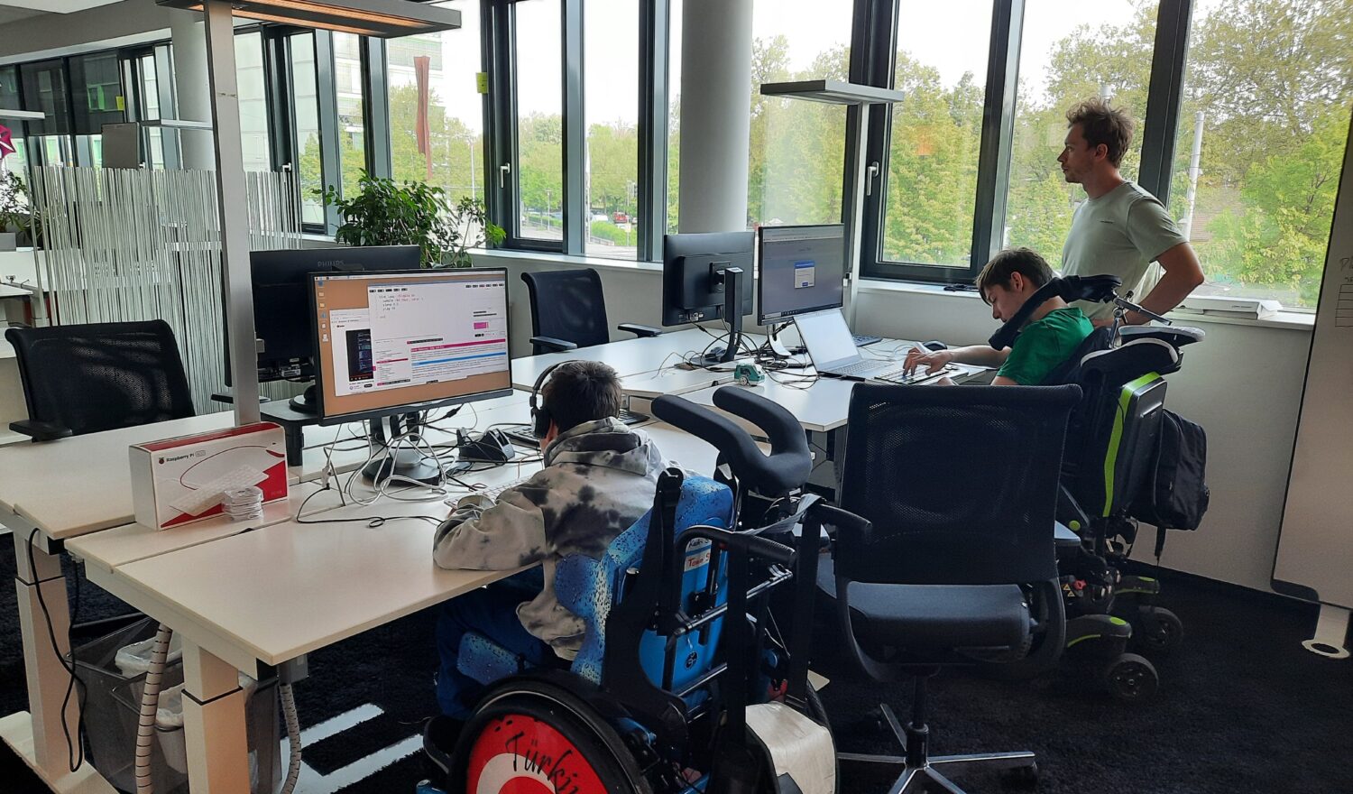 Zwei Kinder, im Rollstuhl, sitzen for Bildschirmen in einem Büro. ein Mann steht neben Ihnen.