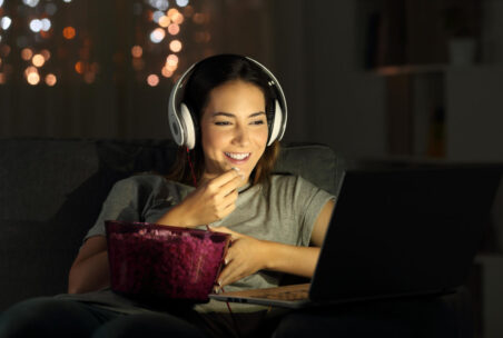 Eine Person mit Kopfhörern und Popcorn-Schüssel in der Hand schaut nachts am Laptop etwas an